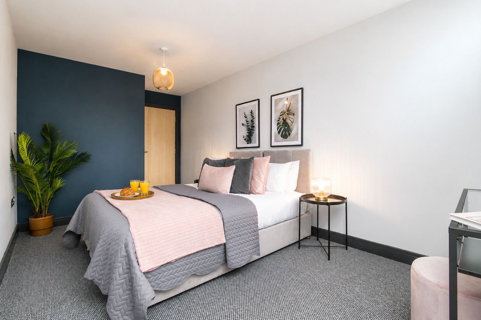 A modern hotel room with a bed, bedside table, potted plant, and artwork on the wall.