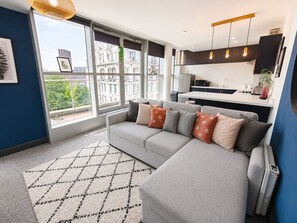 A modern living room with a grey sofa, a patterned rug, and a view of buildings through large windows. - K Suites - 150m to New St Station w/ Balcony (Birmingham)