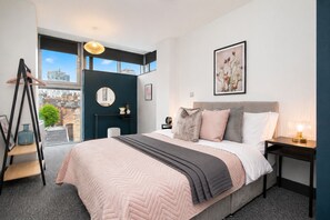 Un dormitorio moderno con una cama grande, un escritorio, una silla, una ventana con vistas a edificios y un espejo redondo en la pared.