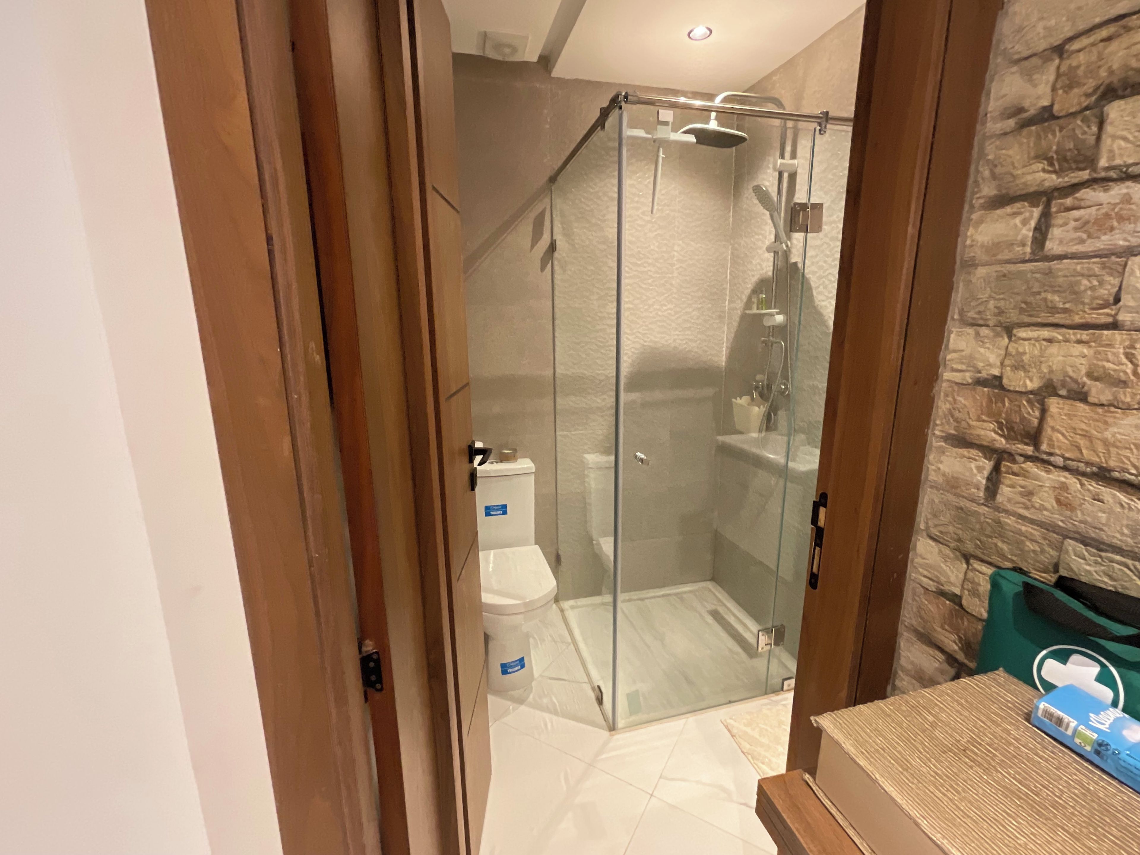 A glass-enclosed shower area with a toilet and a storage bin, a stone-textured wall, and a wooden door frame.