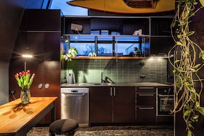A modern kitchen with a built-in dishwasher, a window above the sink, and a small dining area with a wooden table and a vase of flowers. - The Loft - A Design Stay in Battery Point (Battery Point)