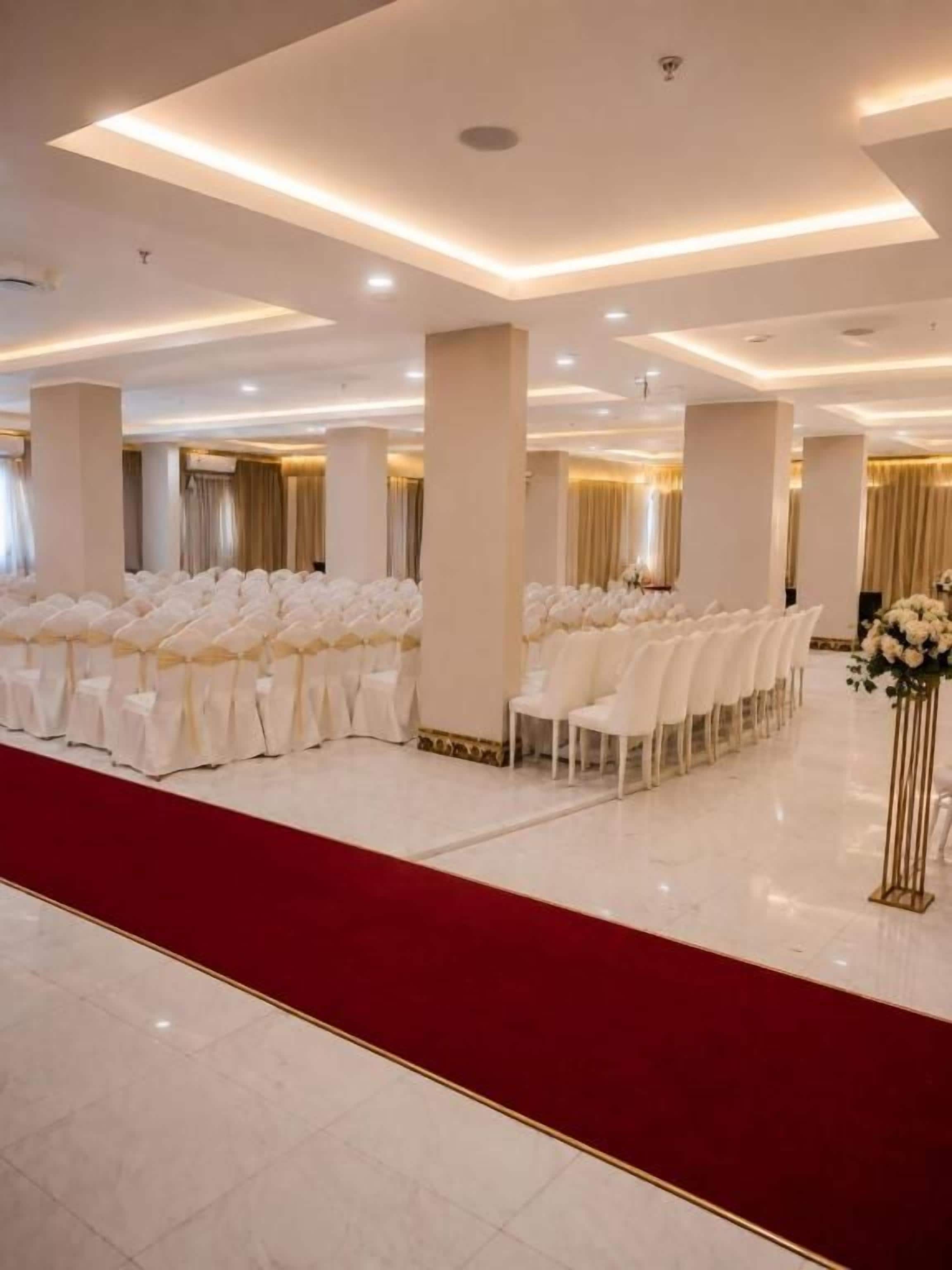 A spacious hall with rows of white chairs, a red carpet, and a decorative floral arrangement.
