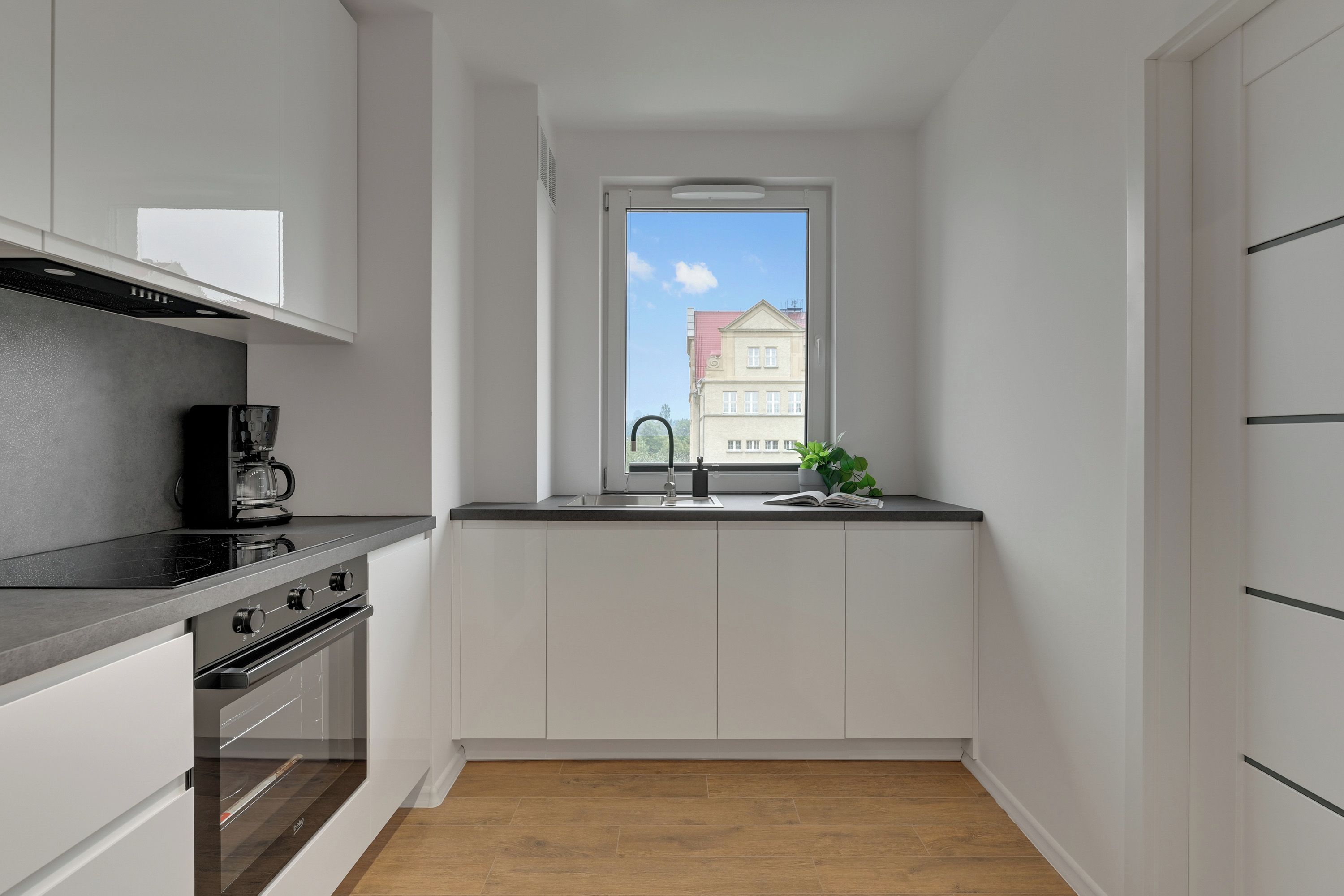 A compact kitchen with a built-in oven, a coffee maker, a window with a view of buildings, and a plant on the counter.