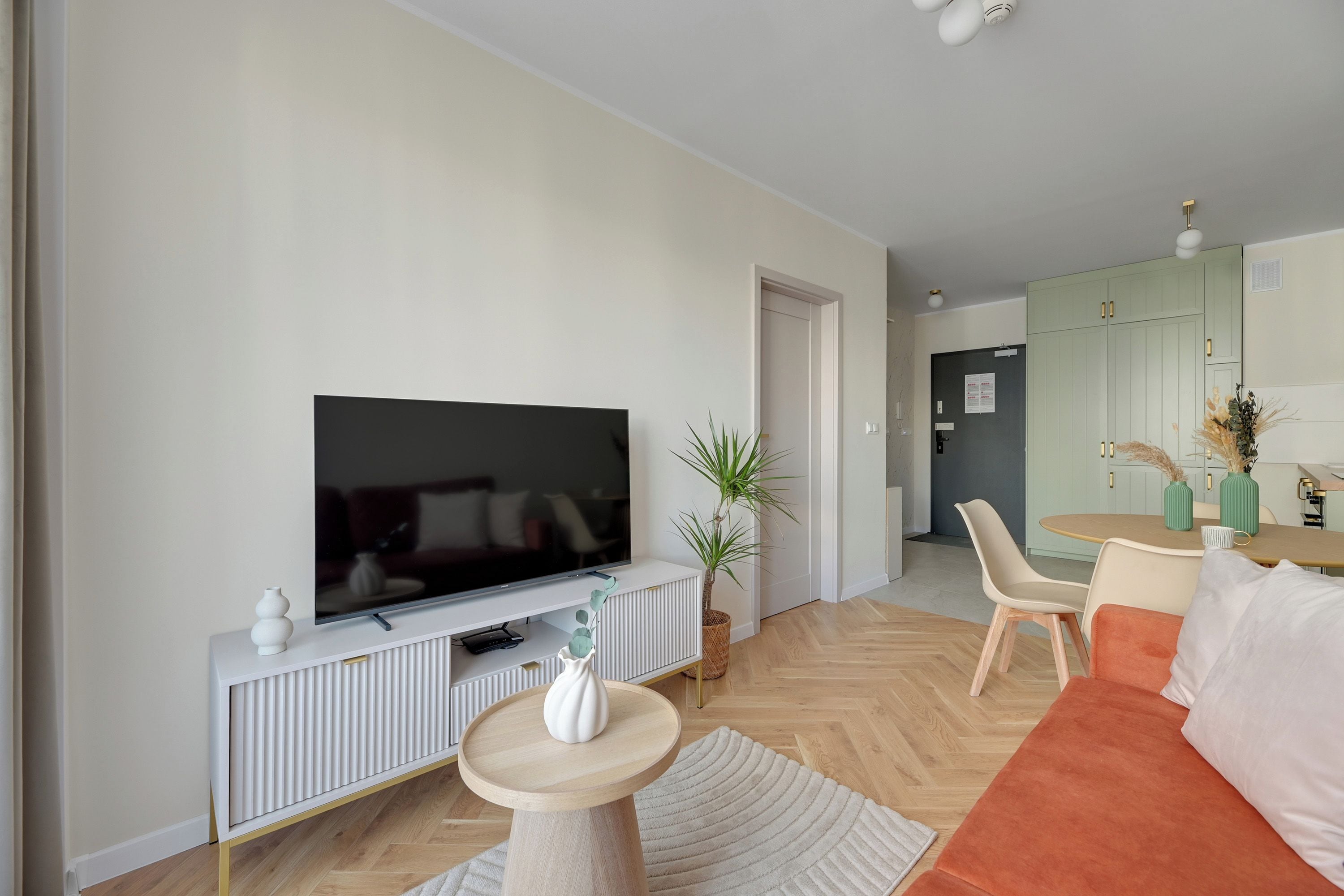 A modern living room with a flat-screen TV, a sofa, a dining table with chairs, and a kitchen area in the background.