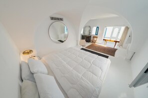 Une chambre avec un grand lit, un miroir rond, une table de chevet et une vue sur un coin repas à travers une fenêtre en arc de cercle.