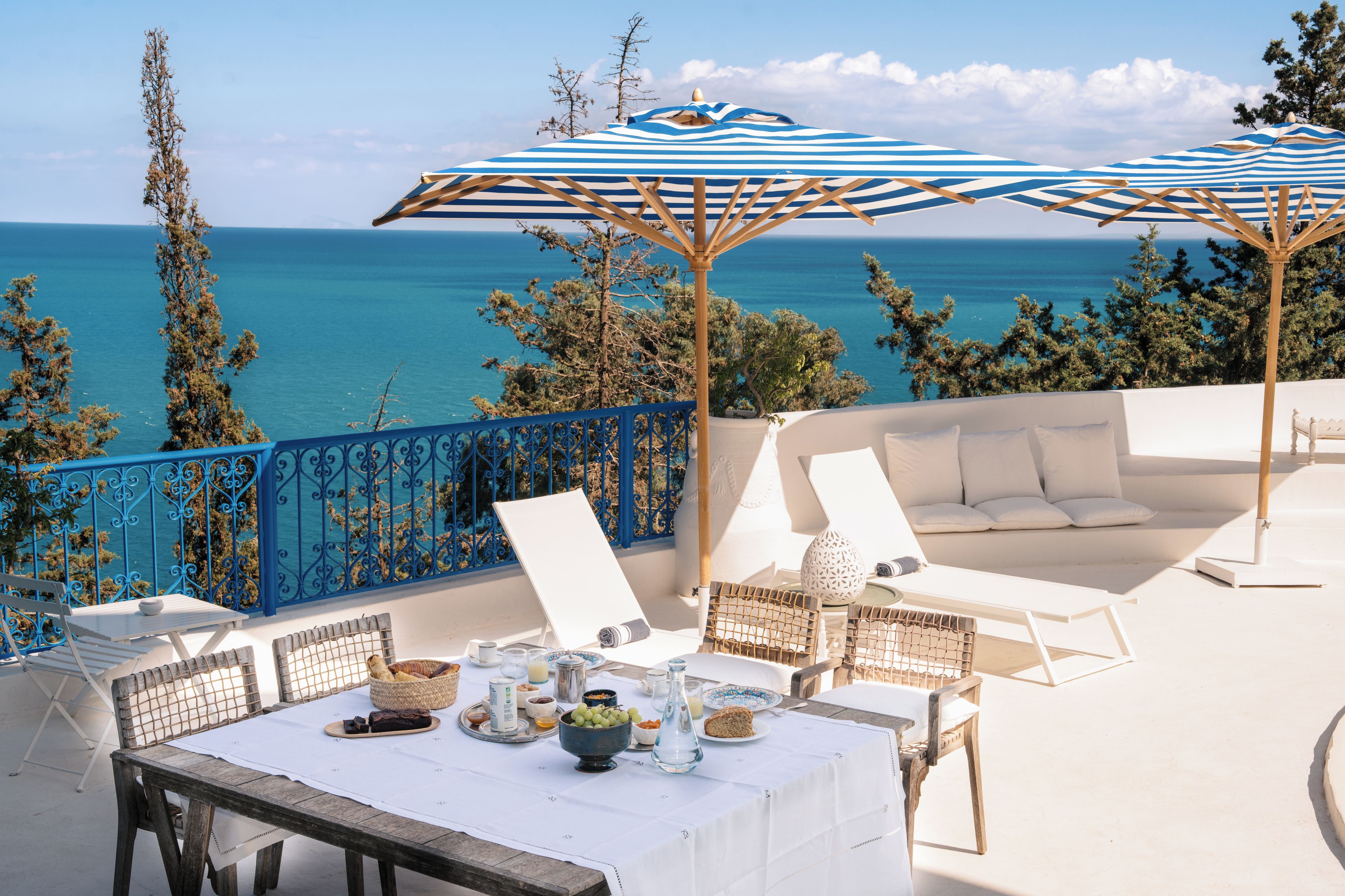 A terrace with a view of the sea, featuring a table with food and drinks, white sofas, and blue umbrellas.