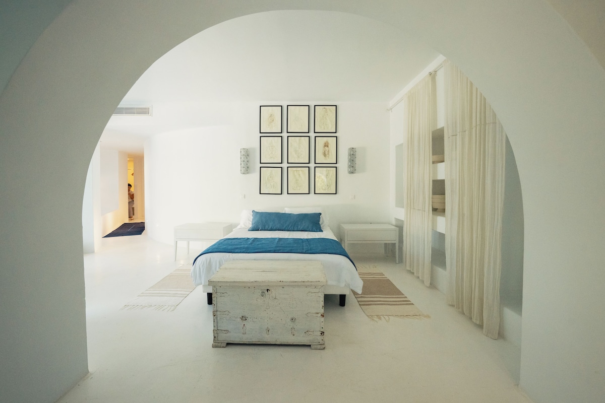 Un dormitorio minimalista con cama, mesita de noche, banco y fotografías enmarcadas en la pared. Cuenta con un ventanal con cortinas.