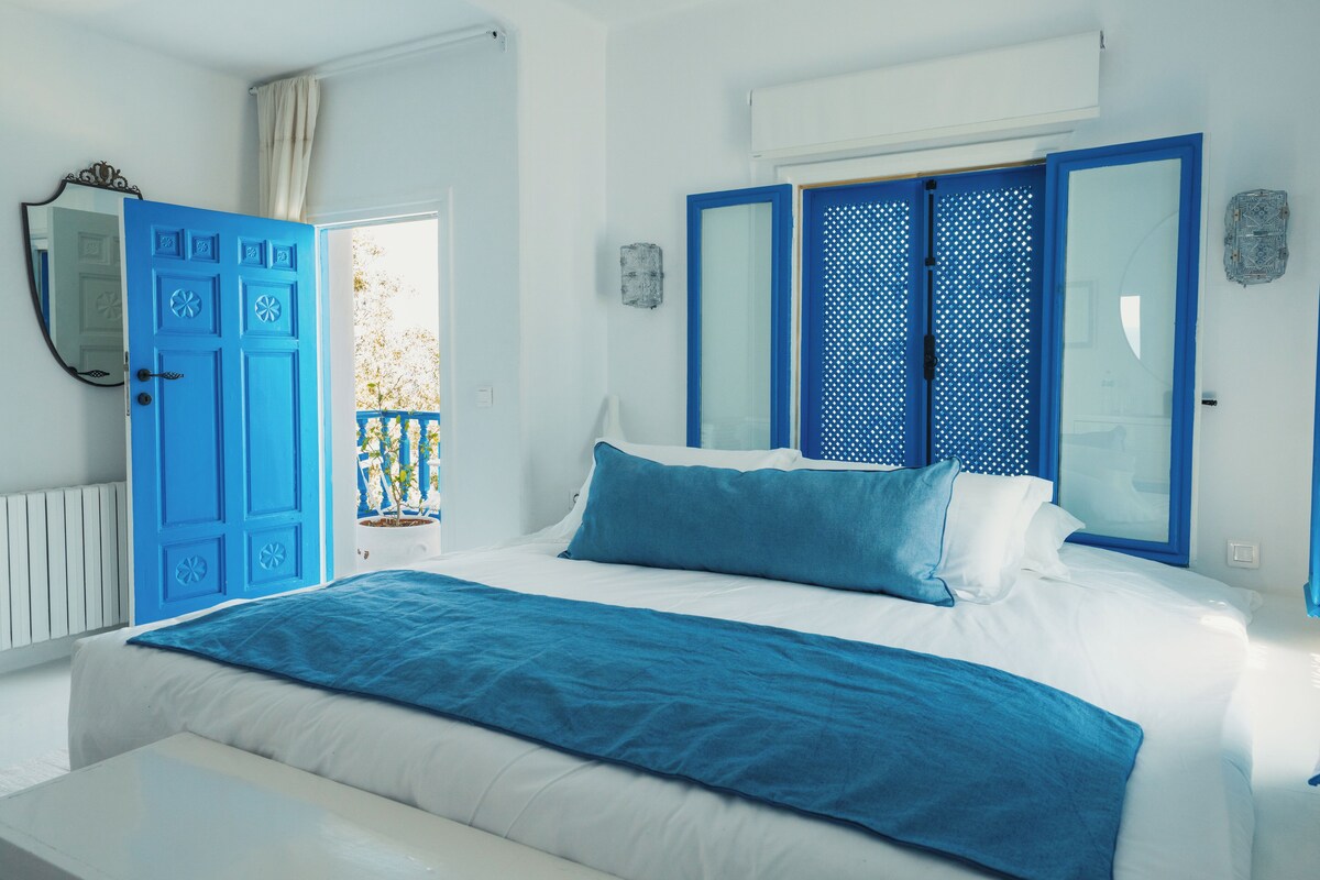 Un dormitorio con cama, ventana con contraventanas azules, radiador blanco y puerta con paneles azules.