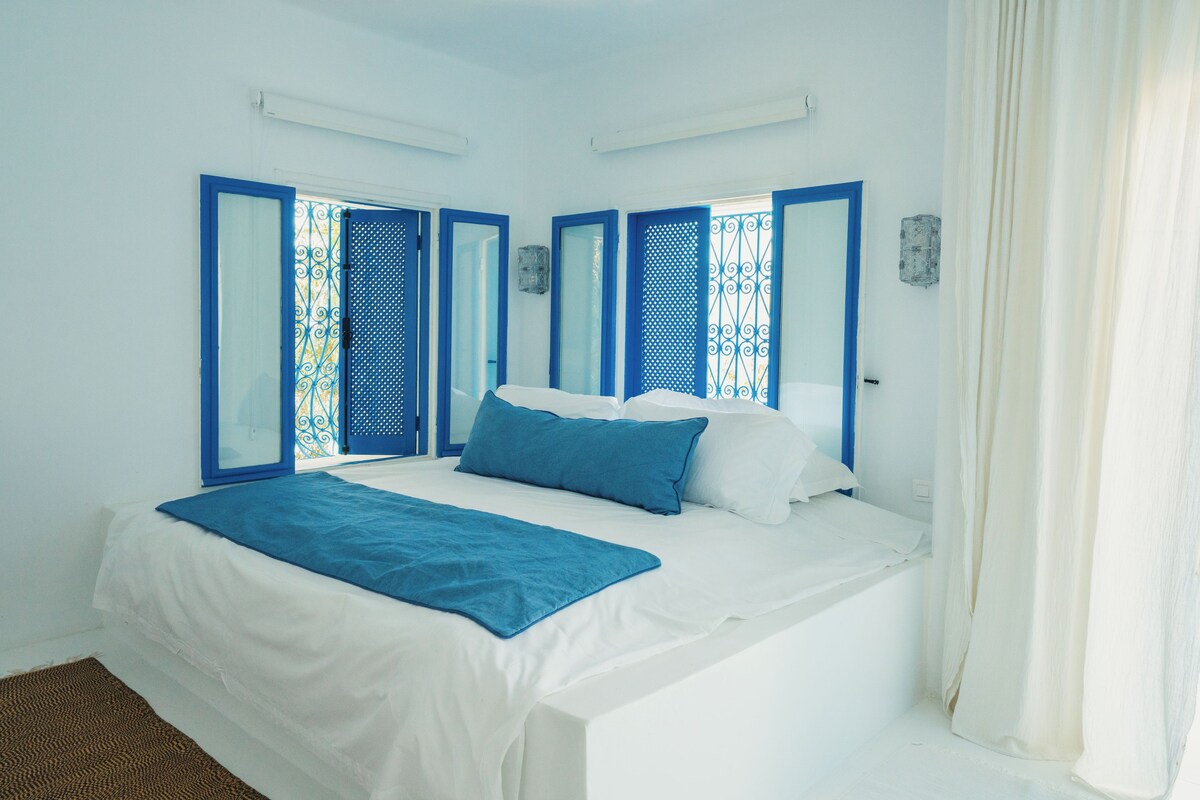 Una habitación con cama, ventana con contraventanas azules y cortina.
