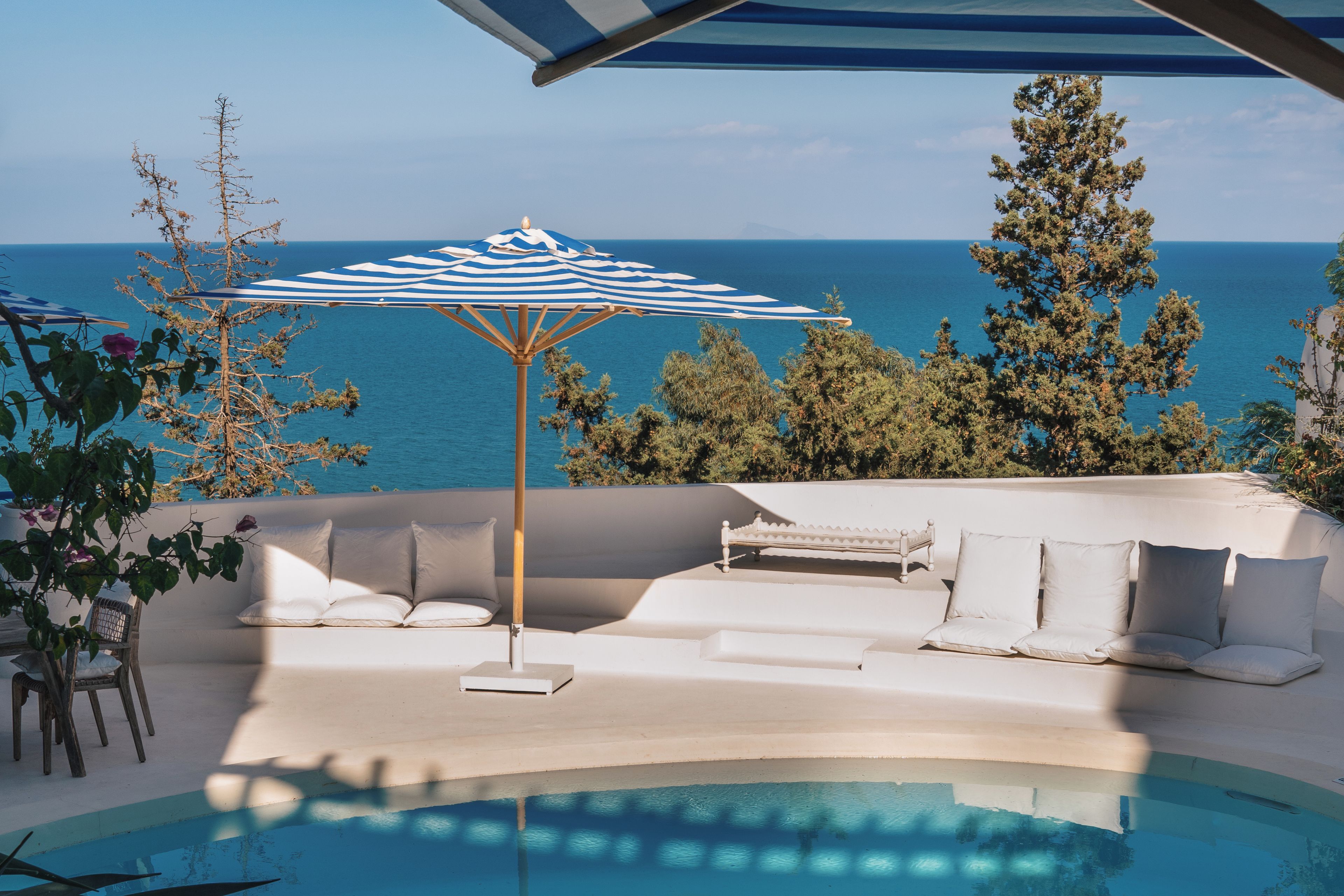 A poolside area with a white outdoor sofa, a striped umbrella, and a view of the sea.