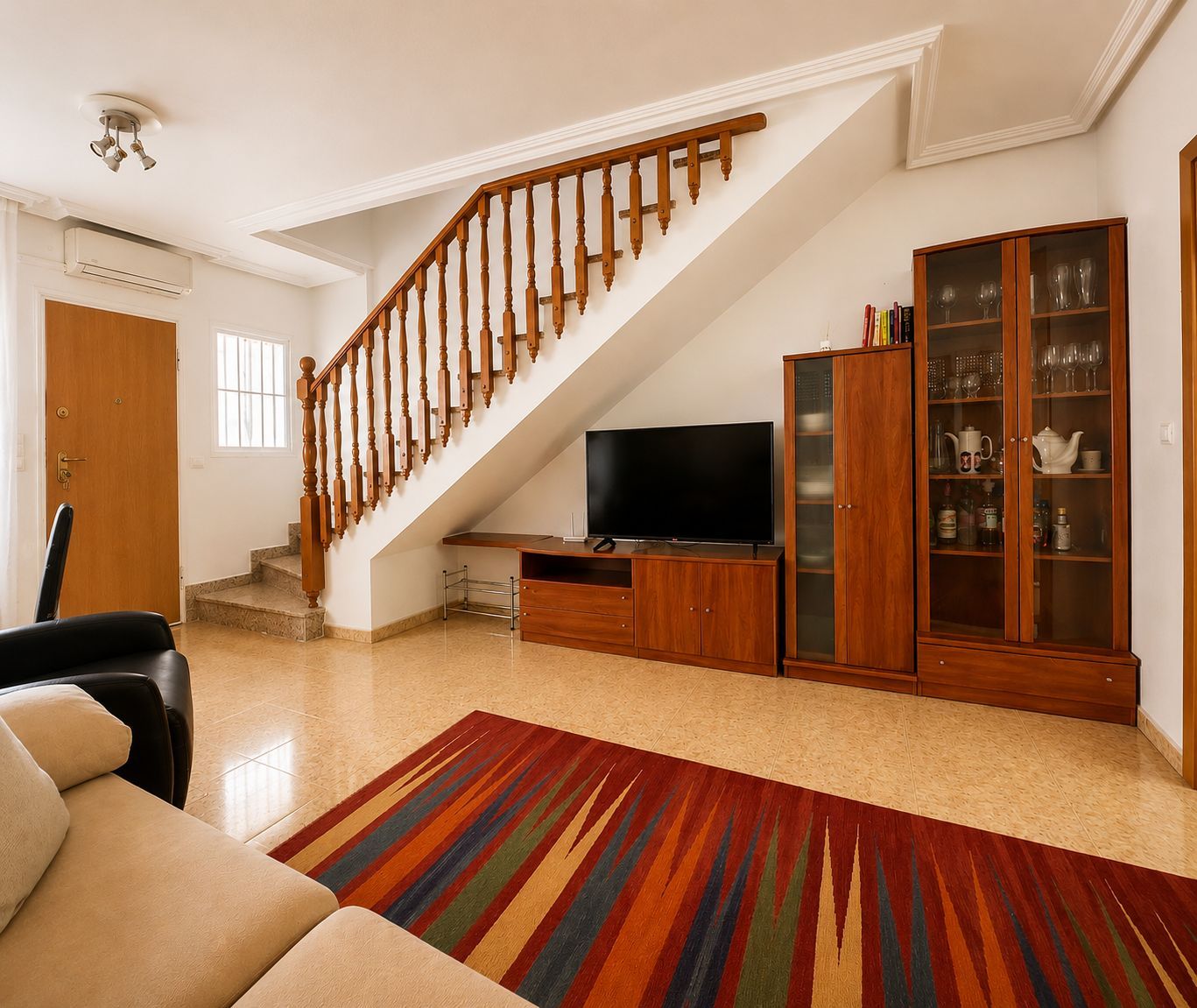 A living room with a wooden staircase, a flat-screen TV, a cabinet with glass doors, and a colorful rug.