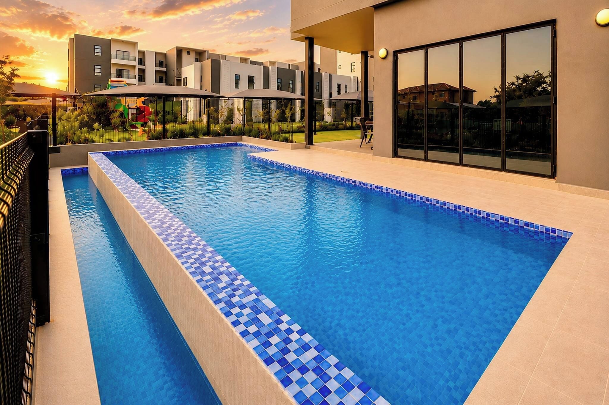 A rectangular swimming pool with blue-tiled edges, surrounded by a modern building with large windows and a flat roof.