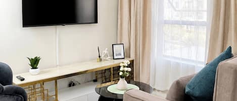 Una sala de estar moderna con un televisor de pantalla plana montado en la pared, un escritorio con una computadora, un sofá, una mesa de centro y una ventana con cortinas.