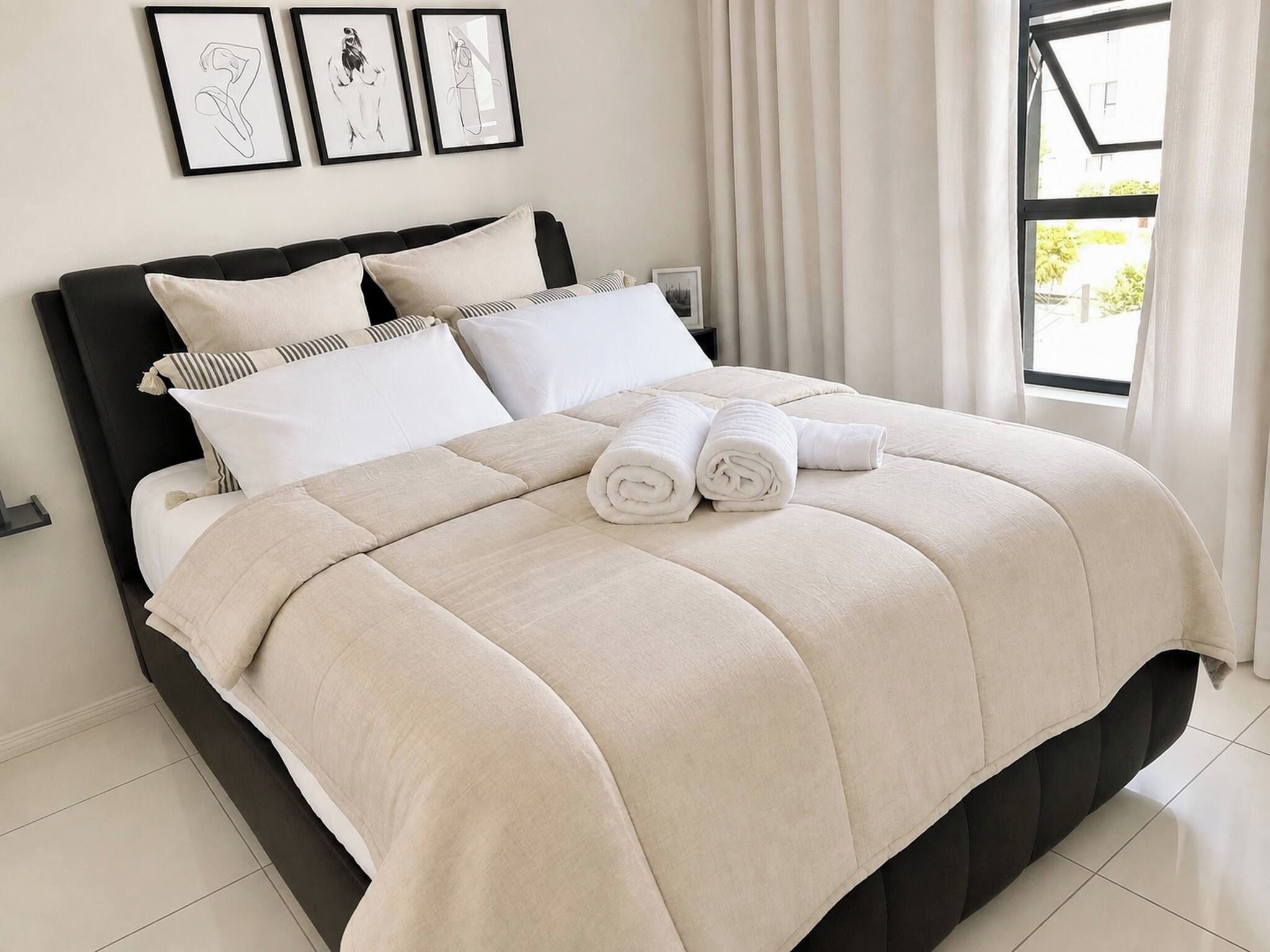 Una cama bien tendida con un edredón color beige, almohadas blancas y toallas enrolladas en blanco.