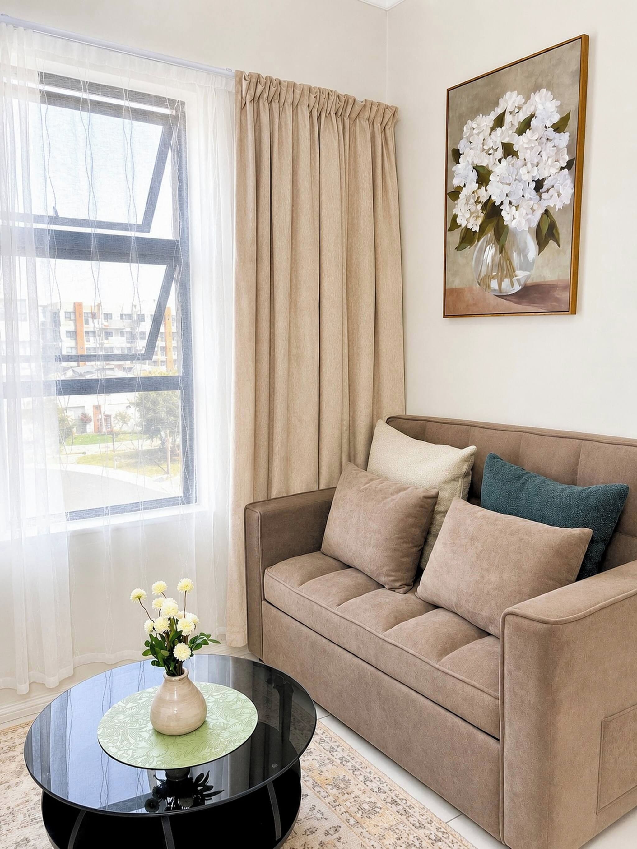 A living room with a sofa, a round coffee table with a vase of flowers, a painting of flowers on the wall, and a view through a window with curtains.