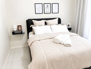 A neatly made bed with a beige quilt, pillows, and rolled towels. There are three framed artworks on the wall above the bed. - Luxury 1BR Suite at 92 on New (Johannesburg)