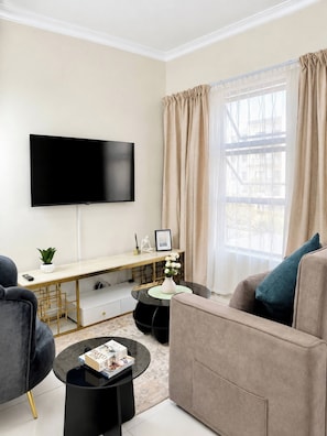 A modern living room with a flat-screen TV mounted on the wall, a desk with a computer, a sofa, a coffee table, and a window with curtains. - Luxury 1BR Suite at 92 on New (Johannesburg)