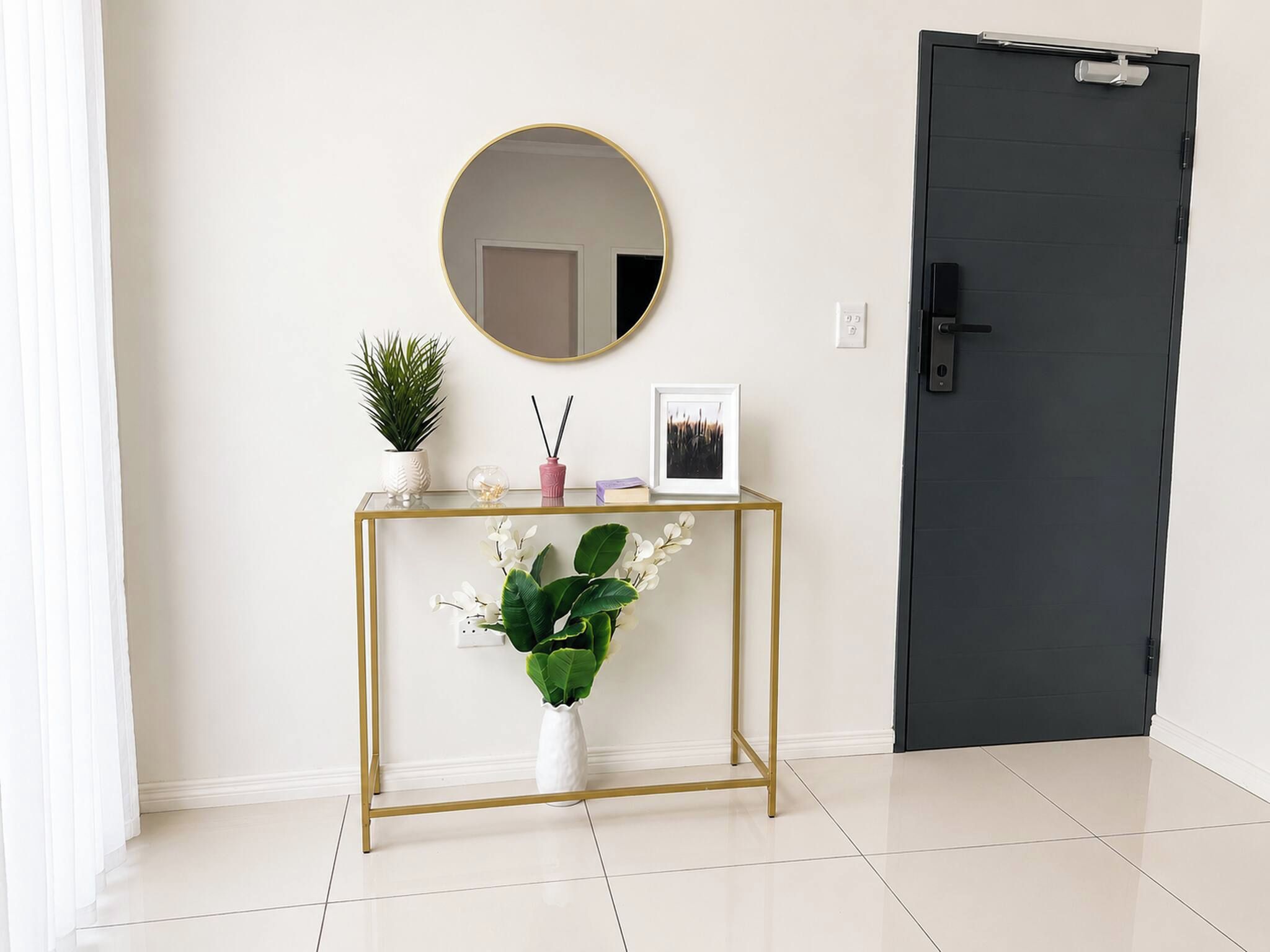 A room with a dark door, a round mirror on a gold-framed console table, a potted plant, and a framed picture.