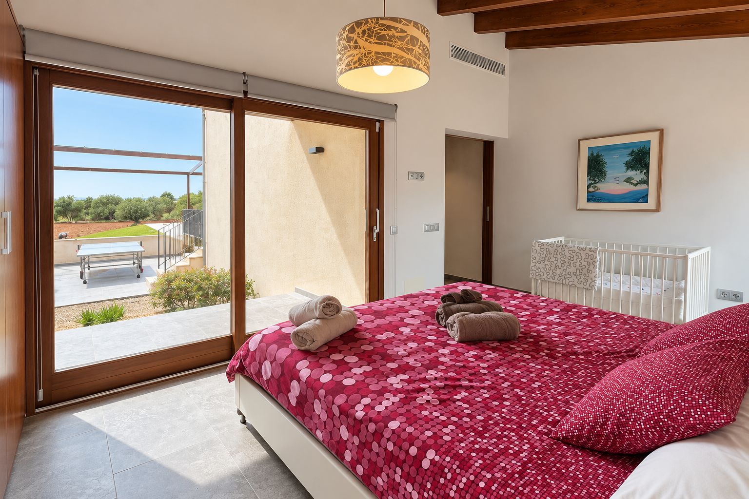 A bedroom with a large bed, a view to the outside, a crib, and a painting on the wall.