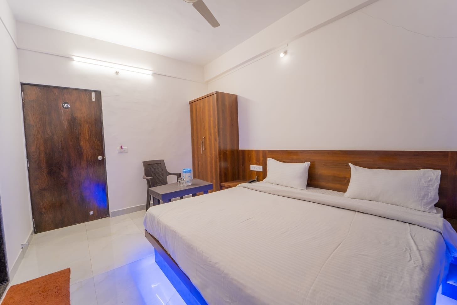 A hotel room with a large bed, a wooden wardrobe, a desk with a chair, and a door marked with the number 103.