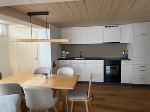 A modern kitchen with white cabinets, a wooden dining table, and grey upholstered chairs. - Grabemattli Appartement/Fewo, Dusche, WC, 1 Schlafraum 2.5-Zimmerwohnung by Interhome (Adelboden)