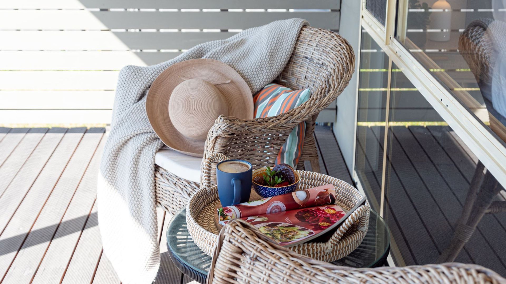 Un panier en osier posé sur une terrasse en bois, avec un chapeau, une tasse et un saladier rempli de fruits.