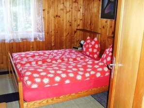 Um quarto de madeira com uma cama, uma janela com cortina translúcida e uma porta.