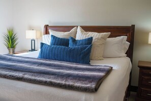Cama bem arrumada com manta escura, cabeceira de madeira e várias almofadas em tons de azul e branco.