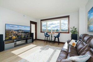 A living room with a large flat-screen TV, a brown leather sofa, two blue chairs, a small round table, and a view of a cityscape through a large window. - Stylish, fully equipped 2-bed / 2-bath condo in Riverstone Village, Coeur d'Alene, with open floor plan, fully equipped kitchen, and secure garage parking. Walkable to nearby dining, shopping, trails, parks and the waterfront. (Coeur d'Alene)
