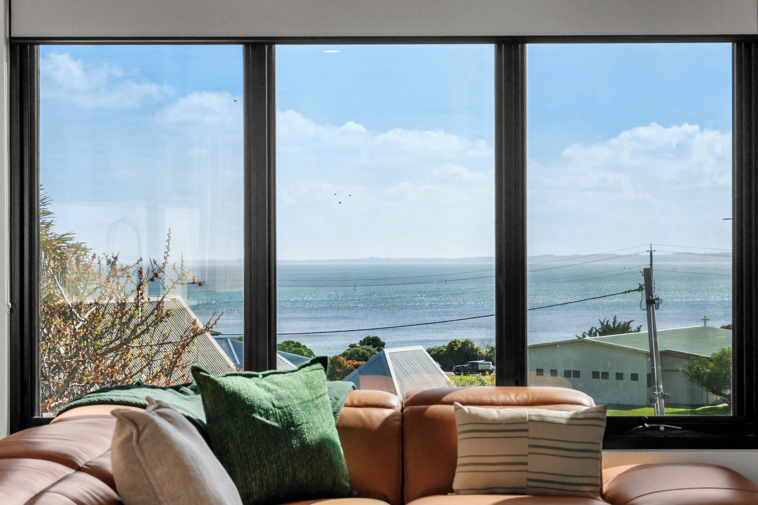 A room with a large window overlooking a body of water, a sofa with cushions, and a view of a distant landscape.
