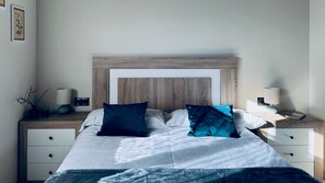 Ein ordentlich bezogenes Bett mit einem Holz-Kopfteil, weißer Bettwäsche und zwei dunklen Kissen. An jeder Seite stehen Nachttische mit Lampen und einer kleinen Pflanze.