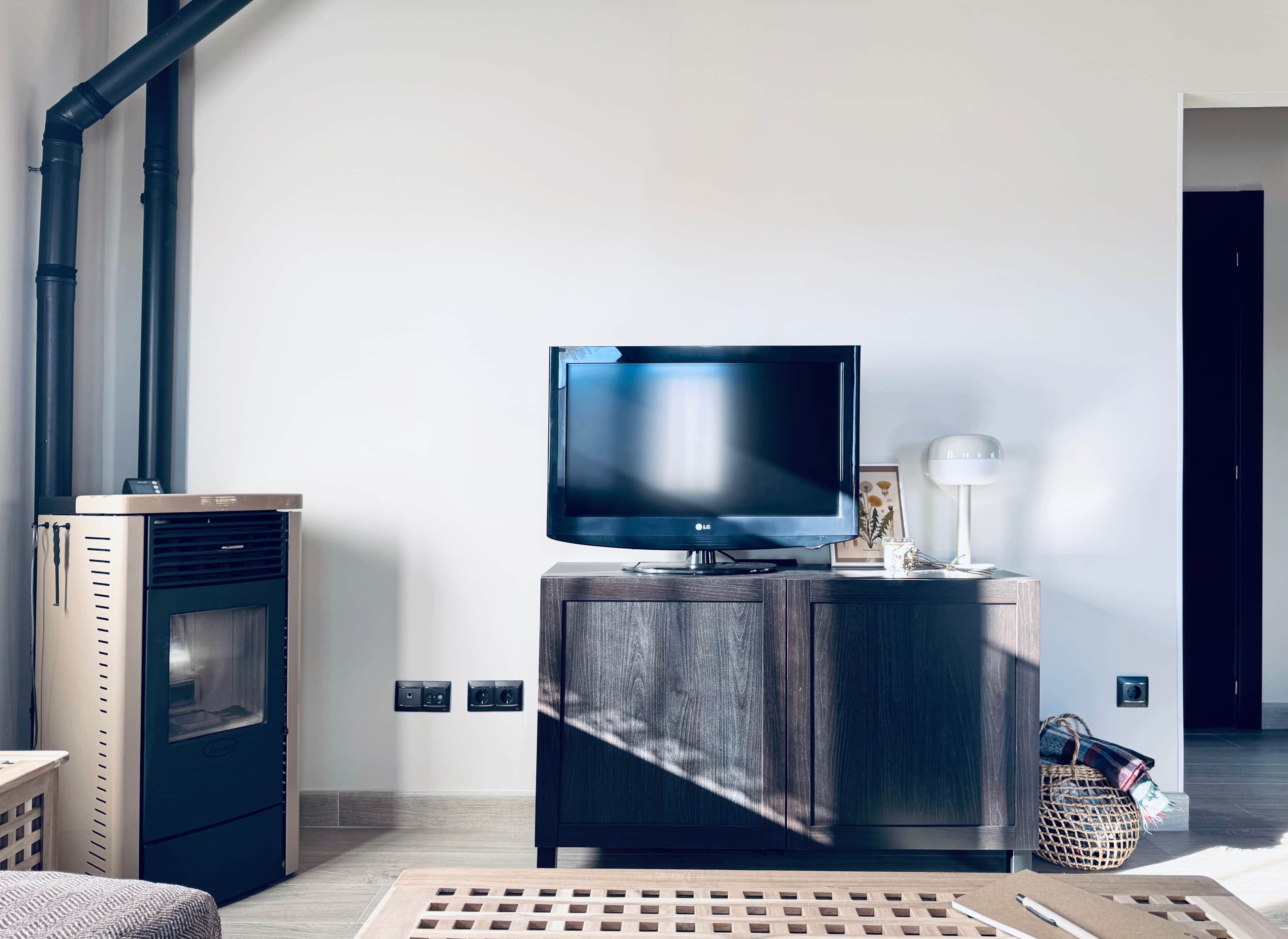 Een moderne woonkamer met een flatscreen televisie op een houten kast, een houtkachel en een witte muur met stopcontacten.