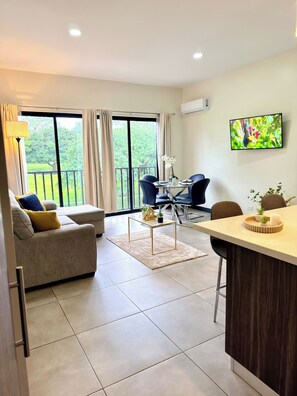 A modern living room with a sofa, a dining table with chairs, a kitchen area with a bar, and a large window with curtains. - Best place near the airport SJO with Air Cond (Alajuela)