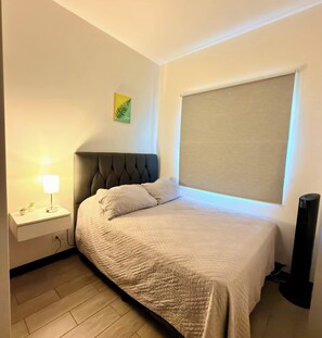 Ein Schlafzimmer mit einem Bett, einem Nachttisch mit Lampe, einem Wandbild und einem großen Fenster mit Jalousie.