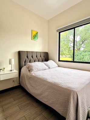 A bedroom with a bed, a bedside table, a lamp, a window with a view of trees, and a wall-mounted picture. - Best place near the airport SJO with Air Cond (Alajuela)