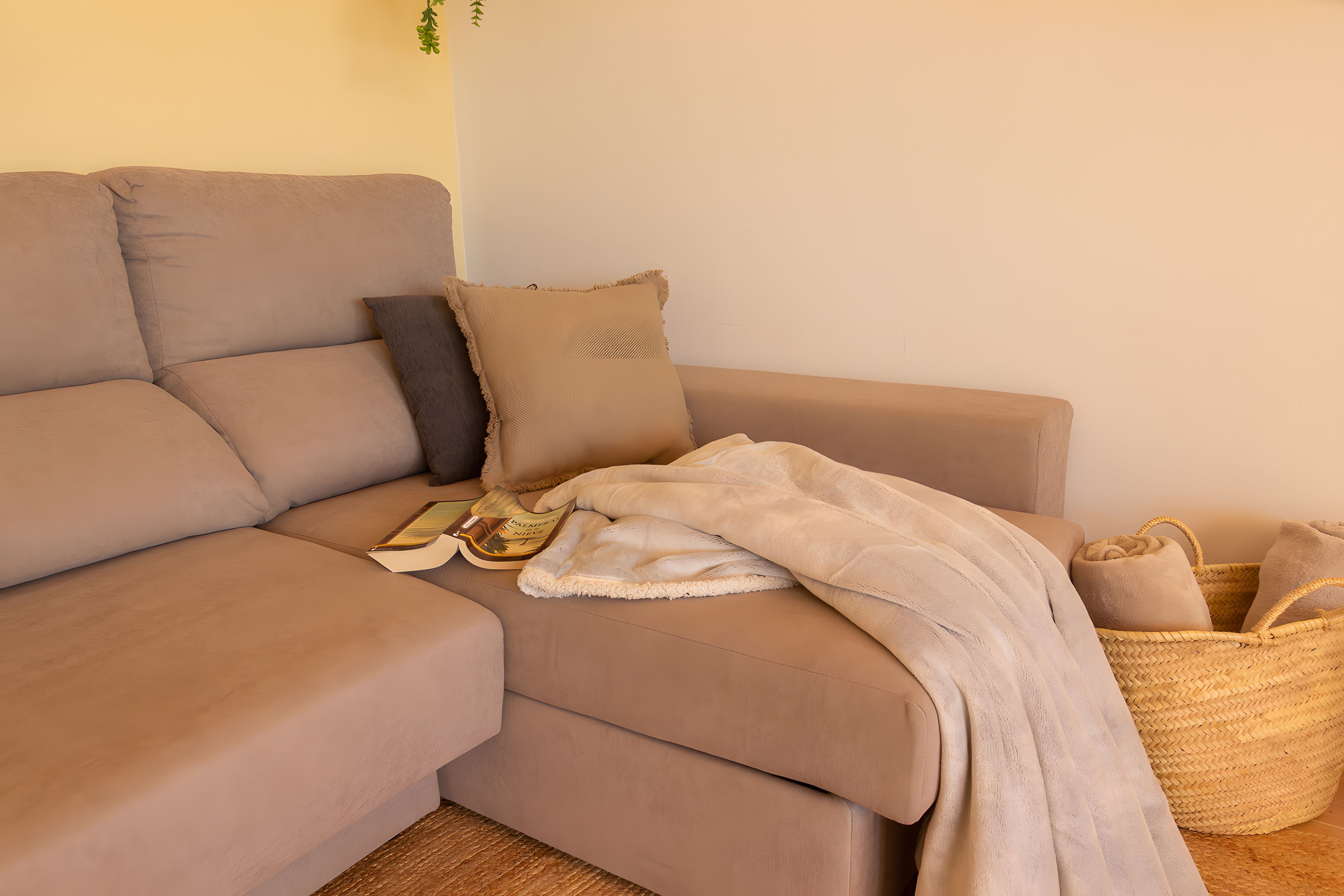 Un sofá beige con una manta, almohadas, un libro y una cesta de mimbre con toallas.