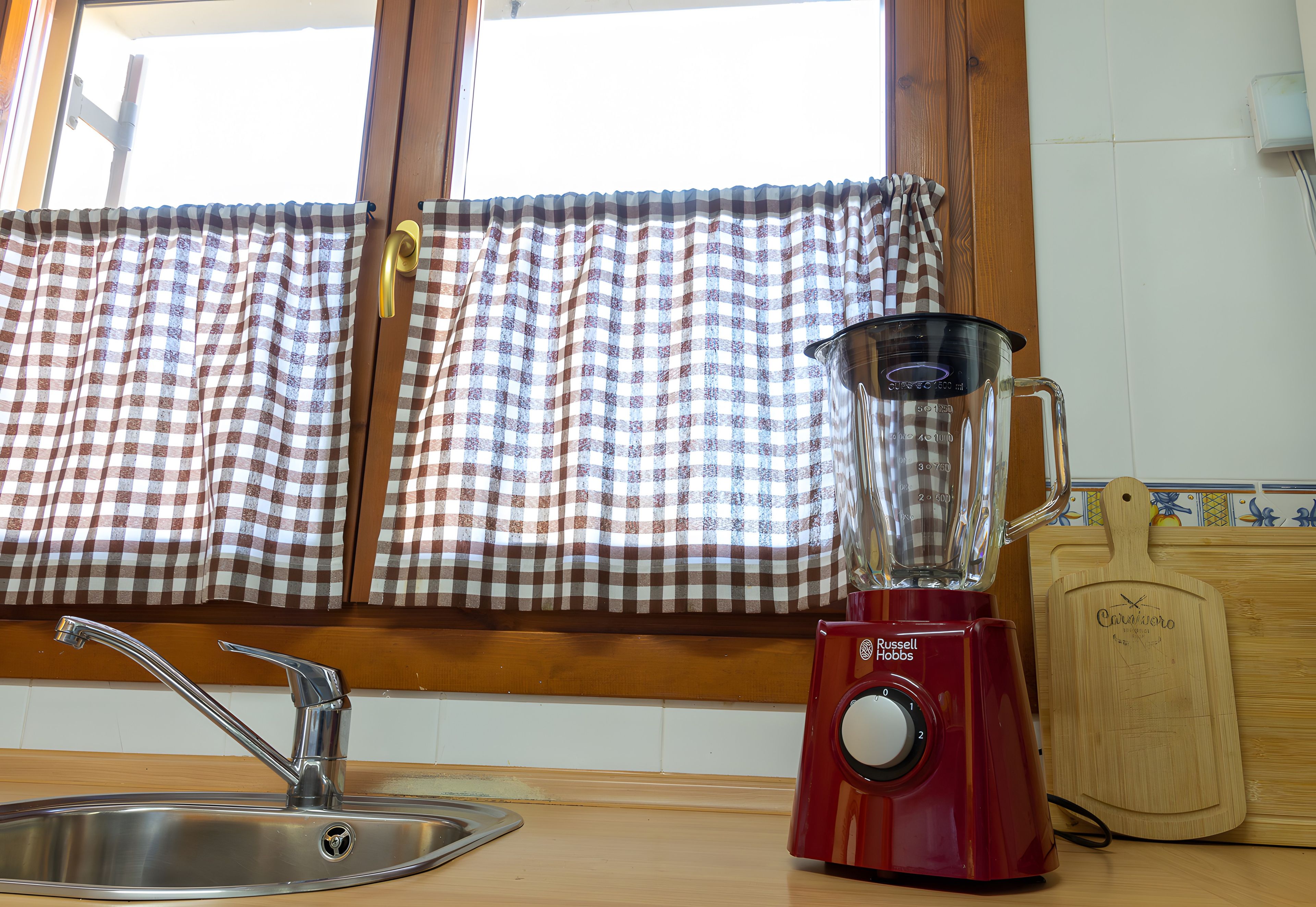 Una cocina con una licuadora roja, un lavamanos de acero inoxidable y una ventana con cortinas de cuadros.