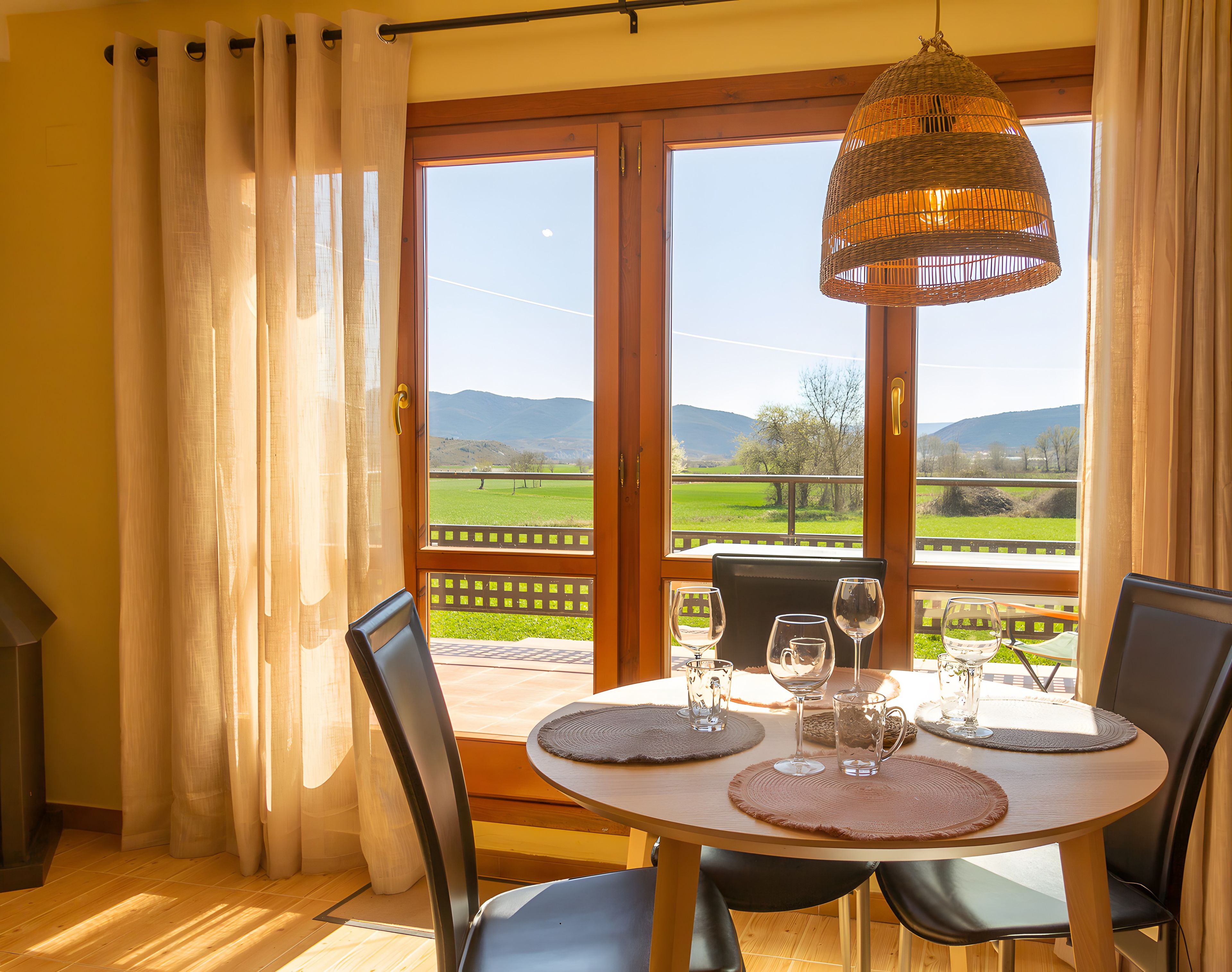 Een eetruimte met een tafel gedekt voor twee personen, een rieten hanglamp en uitzicht op een groen landschap door een glazen deur.