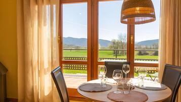Een eetruimte met een tafel gedekt voor twee personen, een rieten hanglamp en uitzicht op een groen landschap door een glazen deur.