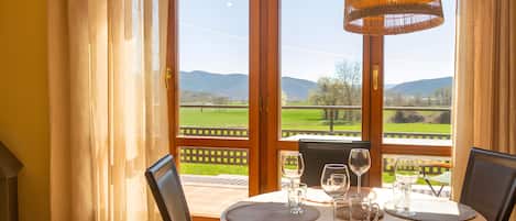 Un coin repas avec une table dressée pour deux personnes, un luminaire suspendu en osier et une vue sur un paysage verdoyant par une porte vitrée.