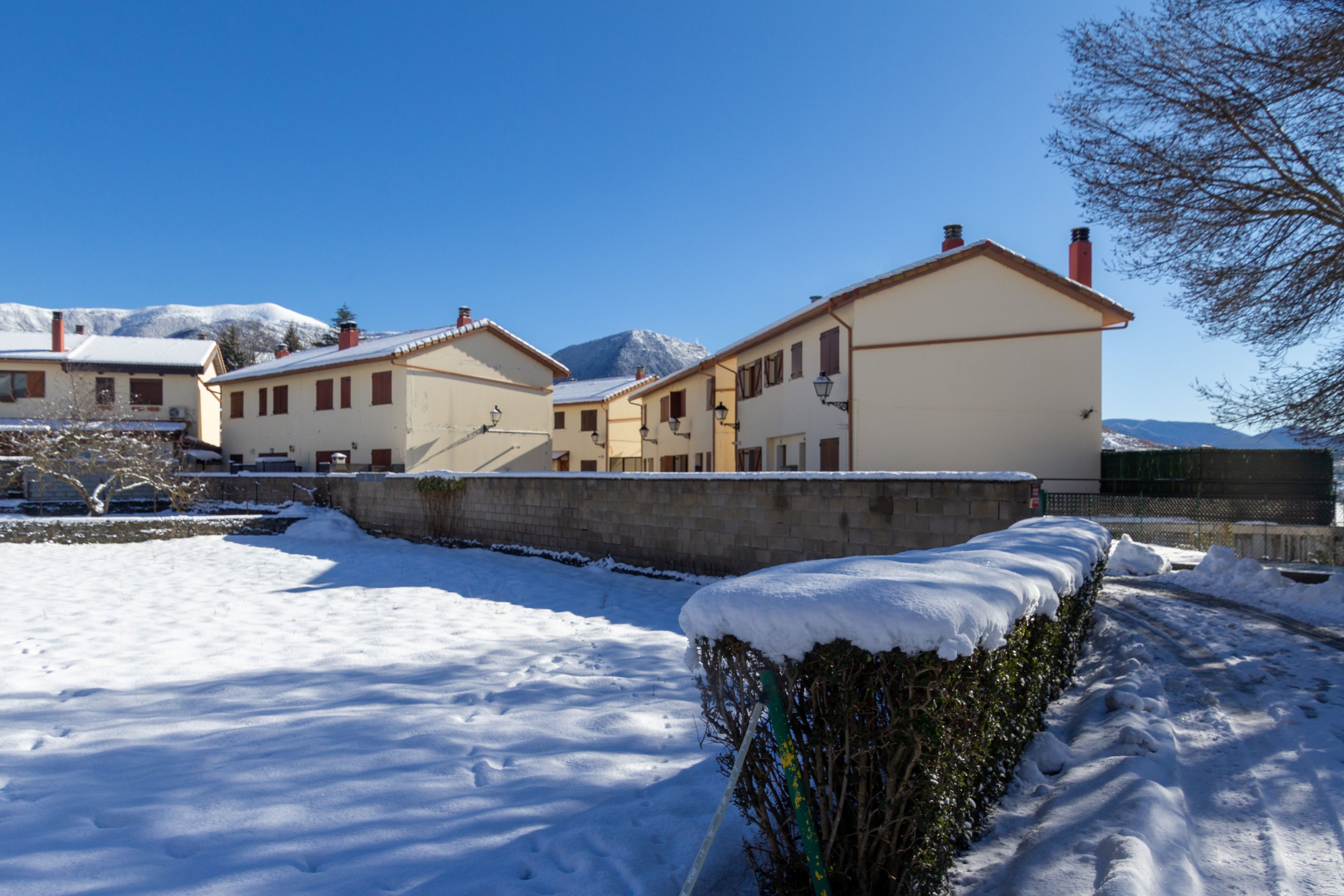 Un patio cubierto de nieve con edificios, un muro de piedra y una montaña al fondo.