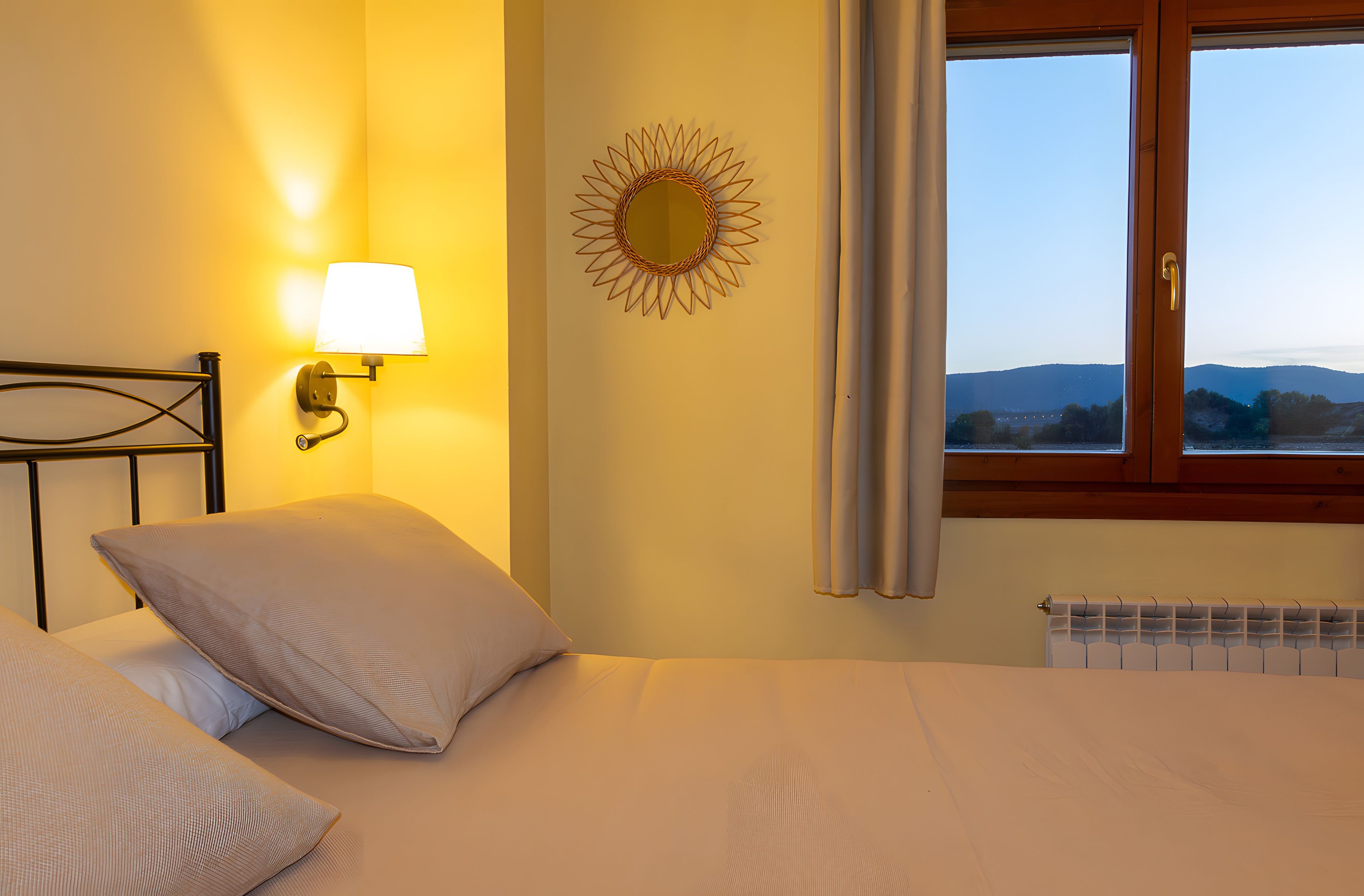 Een slaapkamer met een bed, een wandlamp, een zonnespiegel en een raam met gordijnen.