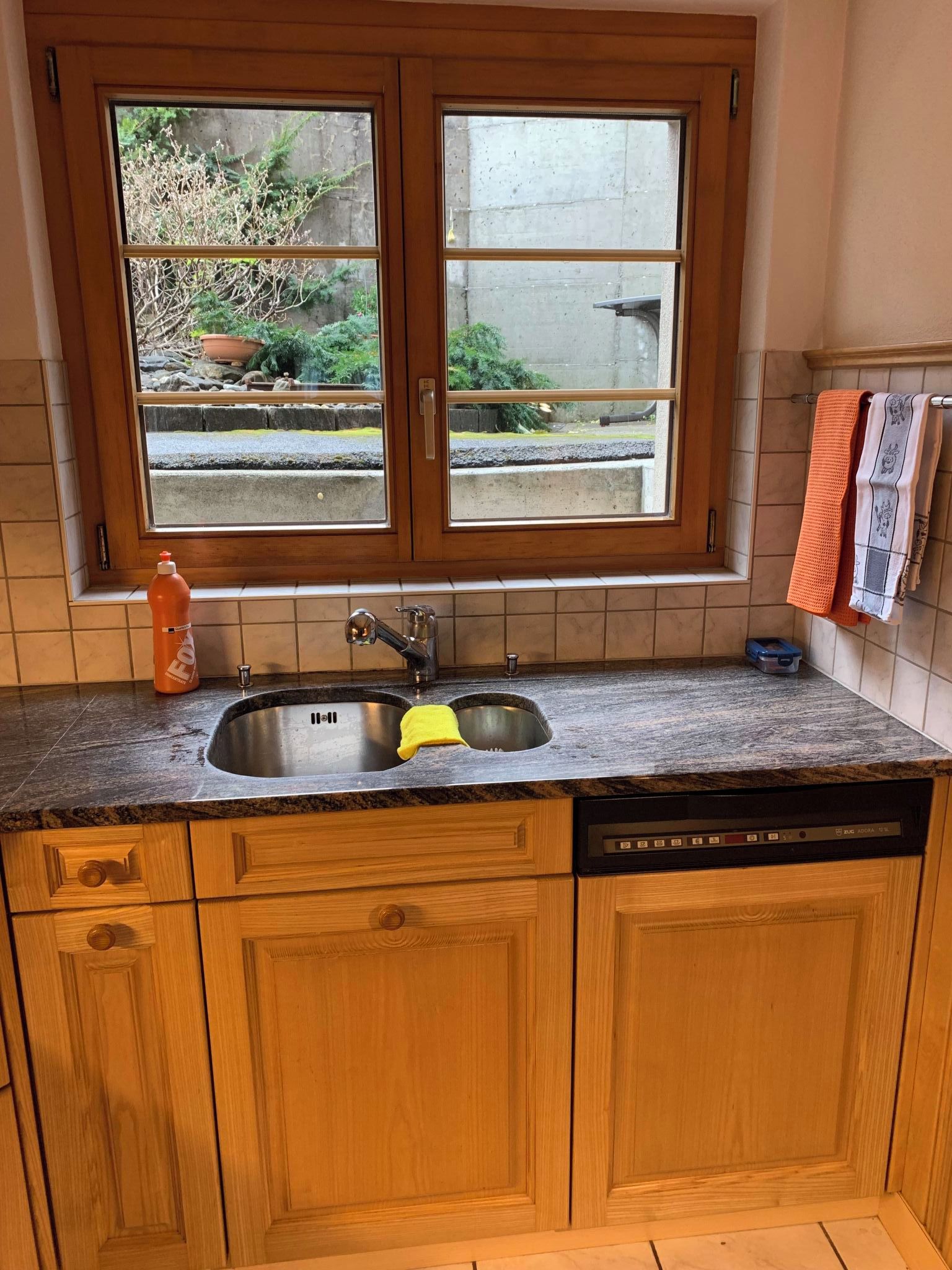 A kitchen with wooden cabinets, a double sink, and a window overlooking a garden.