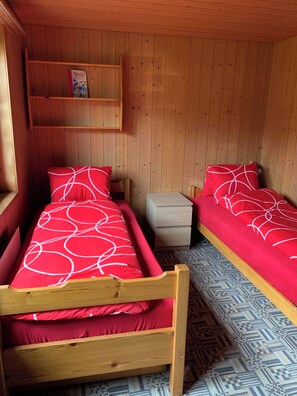 A small, cozy room with two wooden beds, each with red bedding and white patterns. There is a wooden shelf above the beds and a small bedside table. - Alpenglühn Appartement/Fewo, Bad, WC, 3 Schlafräume by Interhome (Adelboden)