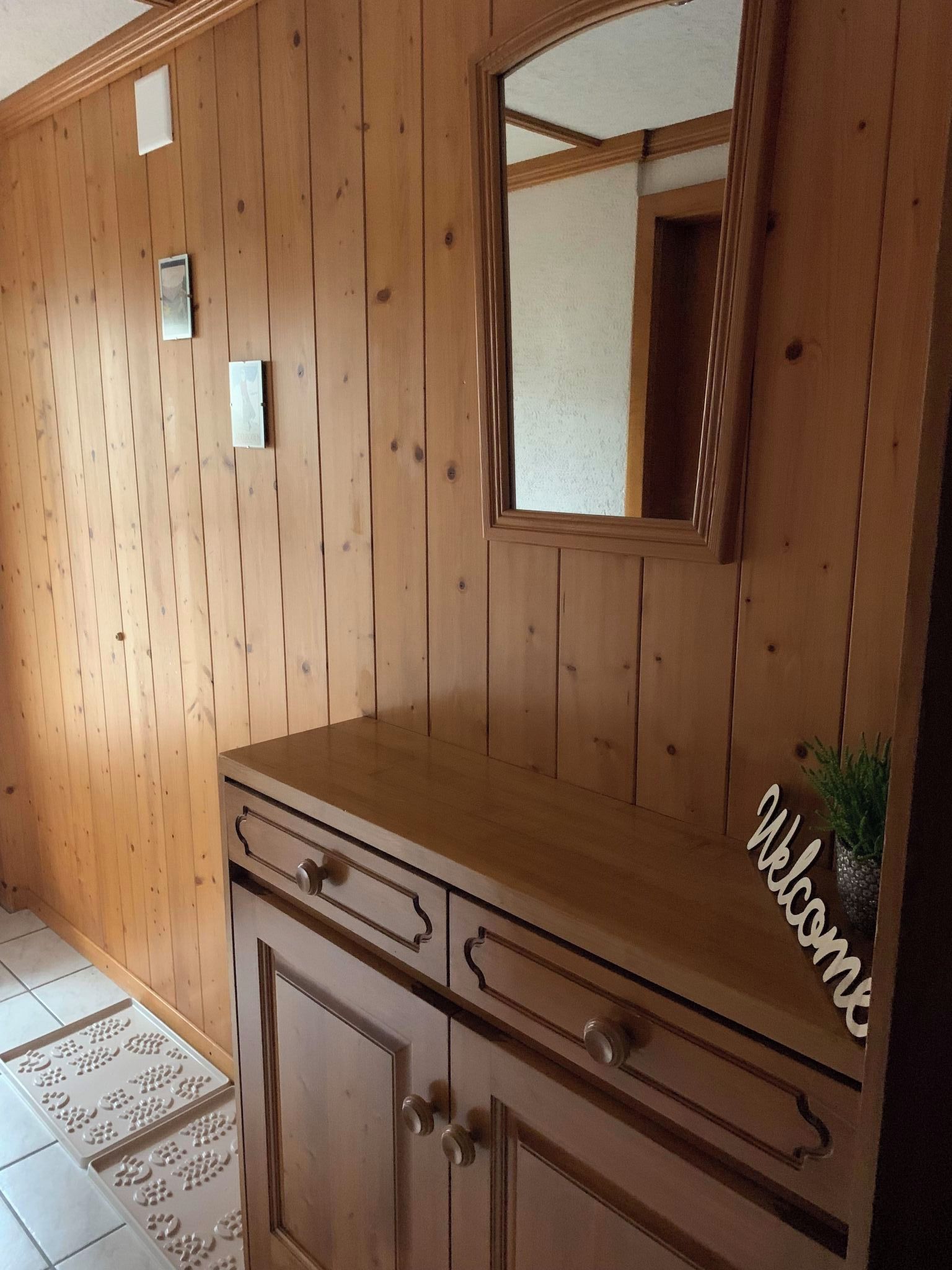 Pared de paneles de madera con un espejo, un mueble con cajones y un cartel de bienvenida.