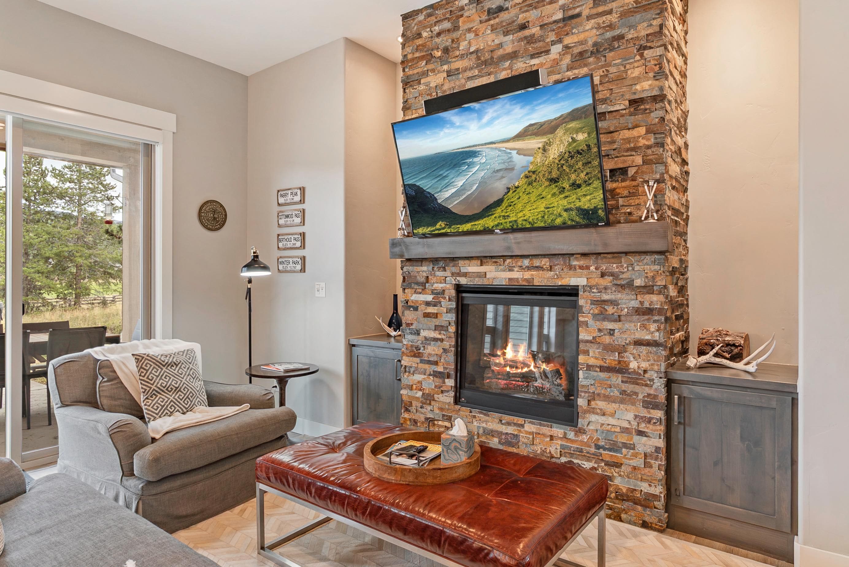 A living room with a stone fireplace, a flat-screen TV mounted above it, and a comfortable seating area with a leather ottoman.