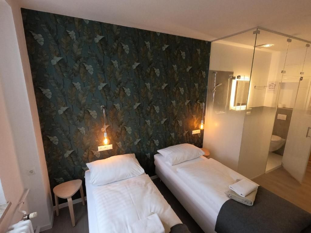 A hotel room with two beds, a small stool, a mirror, and a glass partition separating the bathroom.