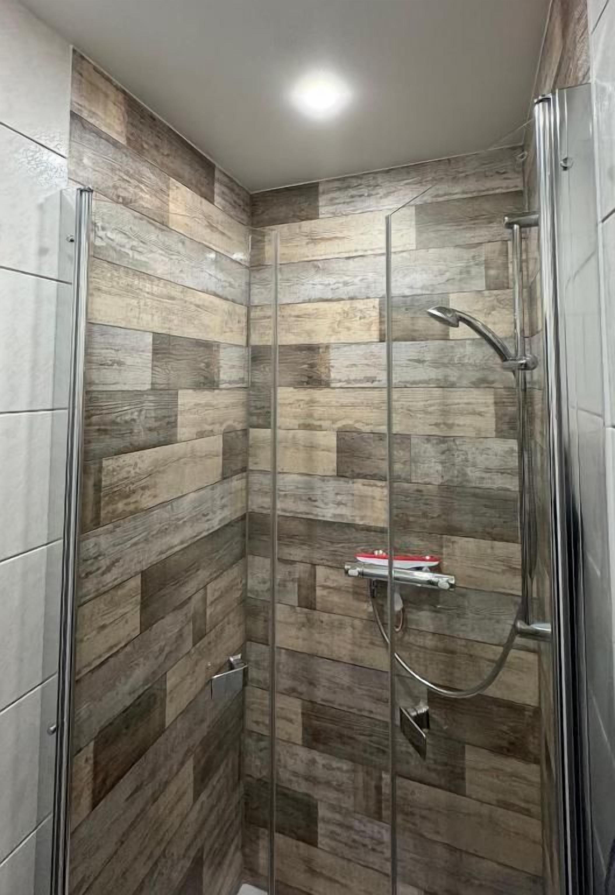 Eine Dusche mit Glastür, Duschkopf aus Metall und einer Wand mit Holzoptik.