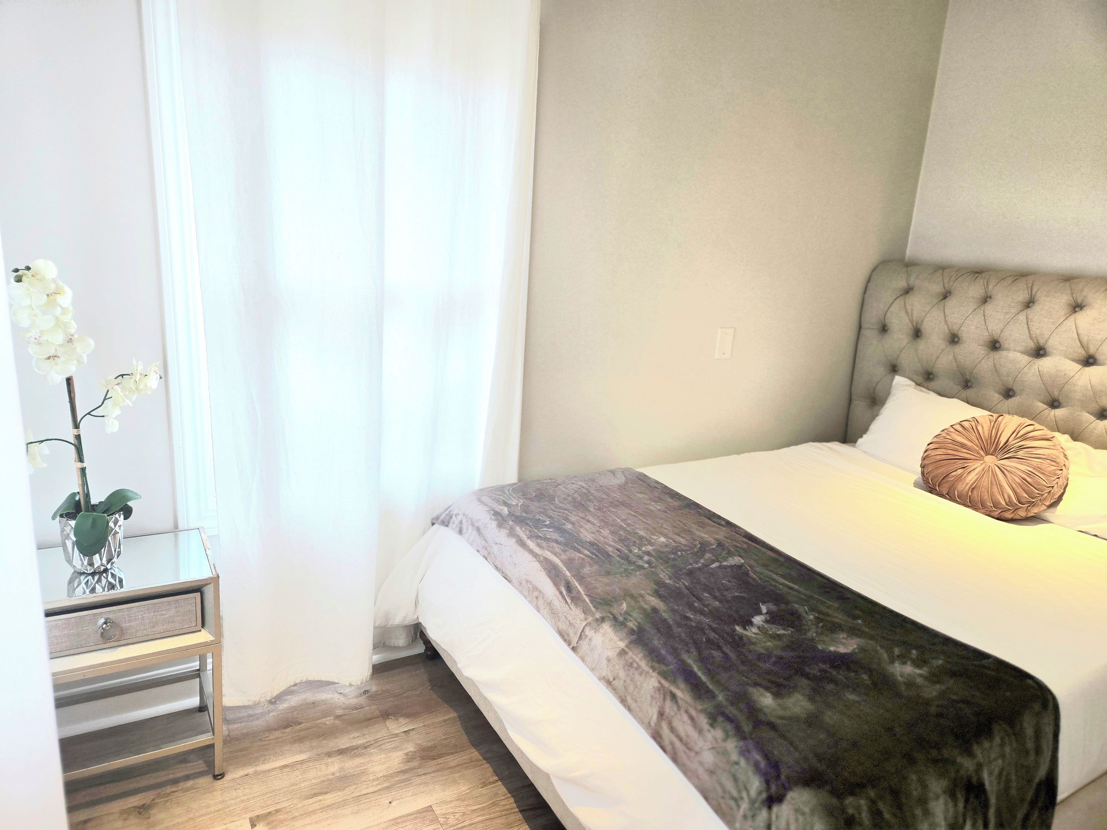 A neatly made bed with a tufted headboard, a bedside table with a drawer, and a potted orchid.