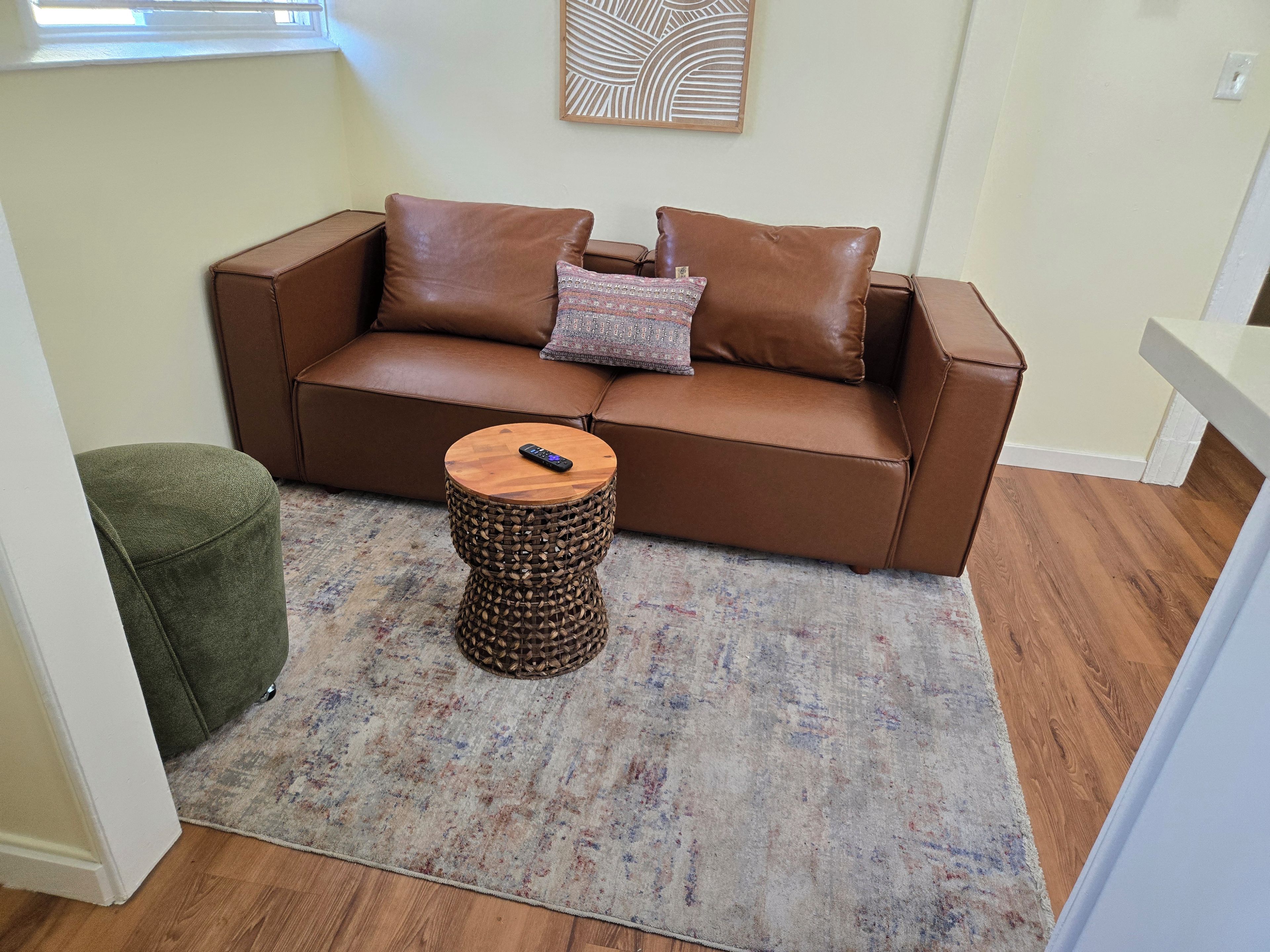 A brown leather sofa with a patterned throw pillow, a small wooden side table with a remote control, and a woven stool.
